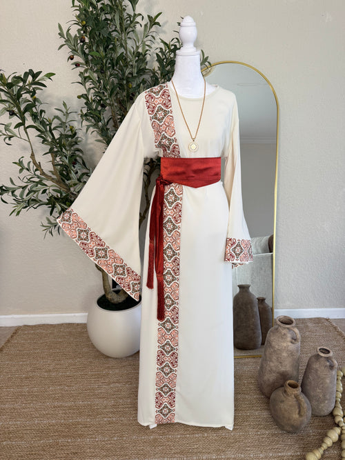 A cream-colored modest dress featuring intricate traditional embroidery along one side and sleeve cuffs, styled with a rich red sash belt, showcasing The Andalusian Abaya.