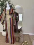 The Desert Bloom Abaya displayed on a mannequin features vibrant vertical stripes in warm tones with a wide sash belt, styled in an elegant, modest fashion setting.