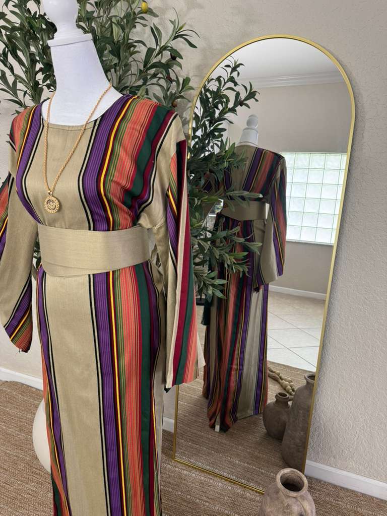 The Desert Bloom Abaya displayed on a mannequin featuring vertical multicolored stripes in earthy tones with a wide beige waist sash, styled with a gold pendant necklace.