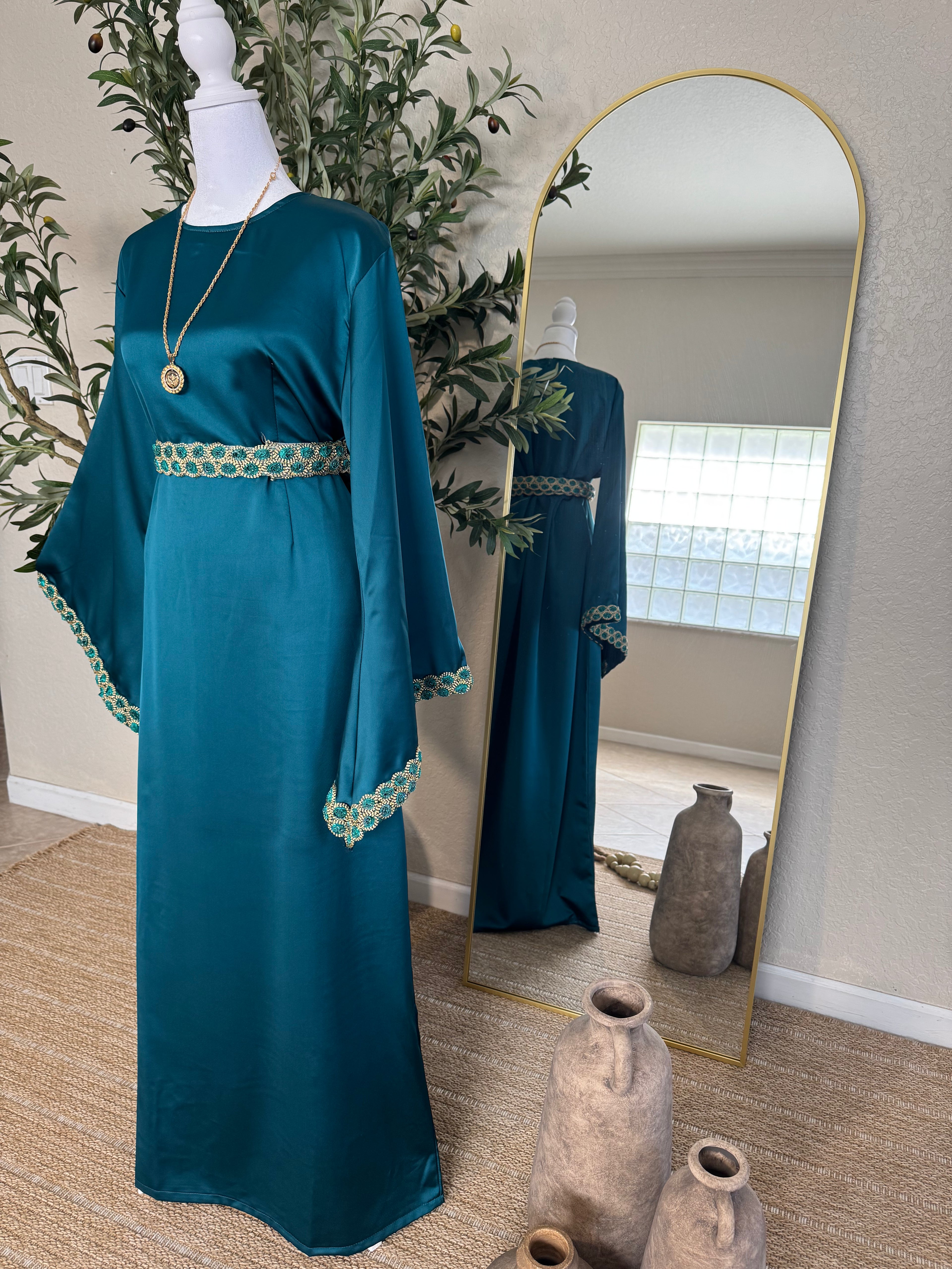 The Emerald Dress displayed on a mannequin next to a tall mirror, featuring teal fabric with gold embroidered trim on the sleeves and waist, styled with a pendant necklace in a minimalist room.