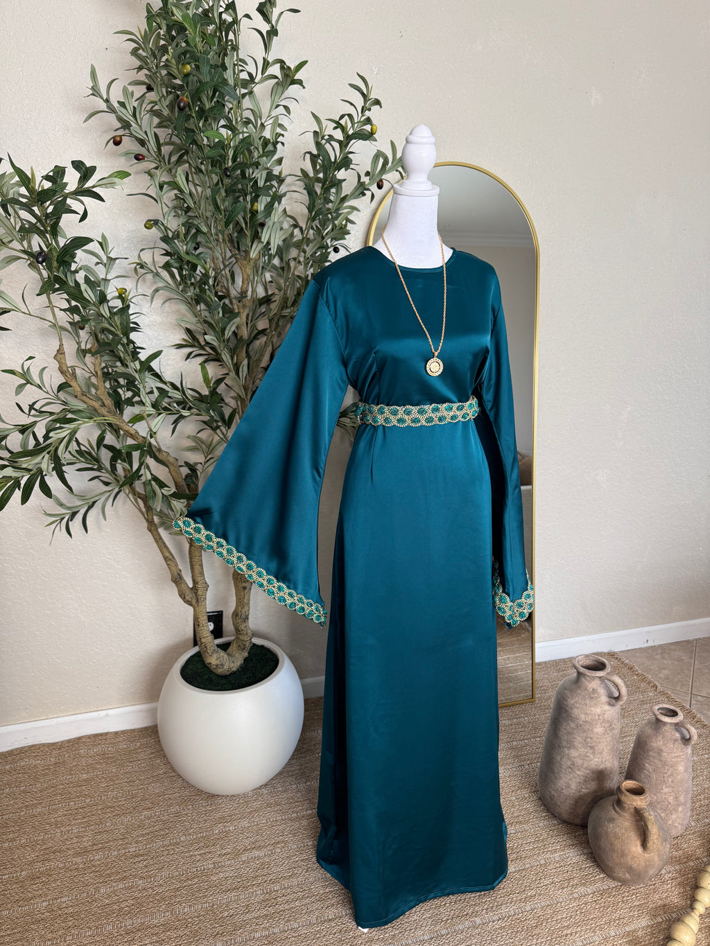 The Emerald Dress in a rich emerald green fabric with wide sleeves and intricate gold embroidery at the cuffs and waist, styled on a mannequin next to a potted olive tree and decorative vases.