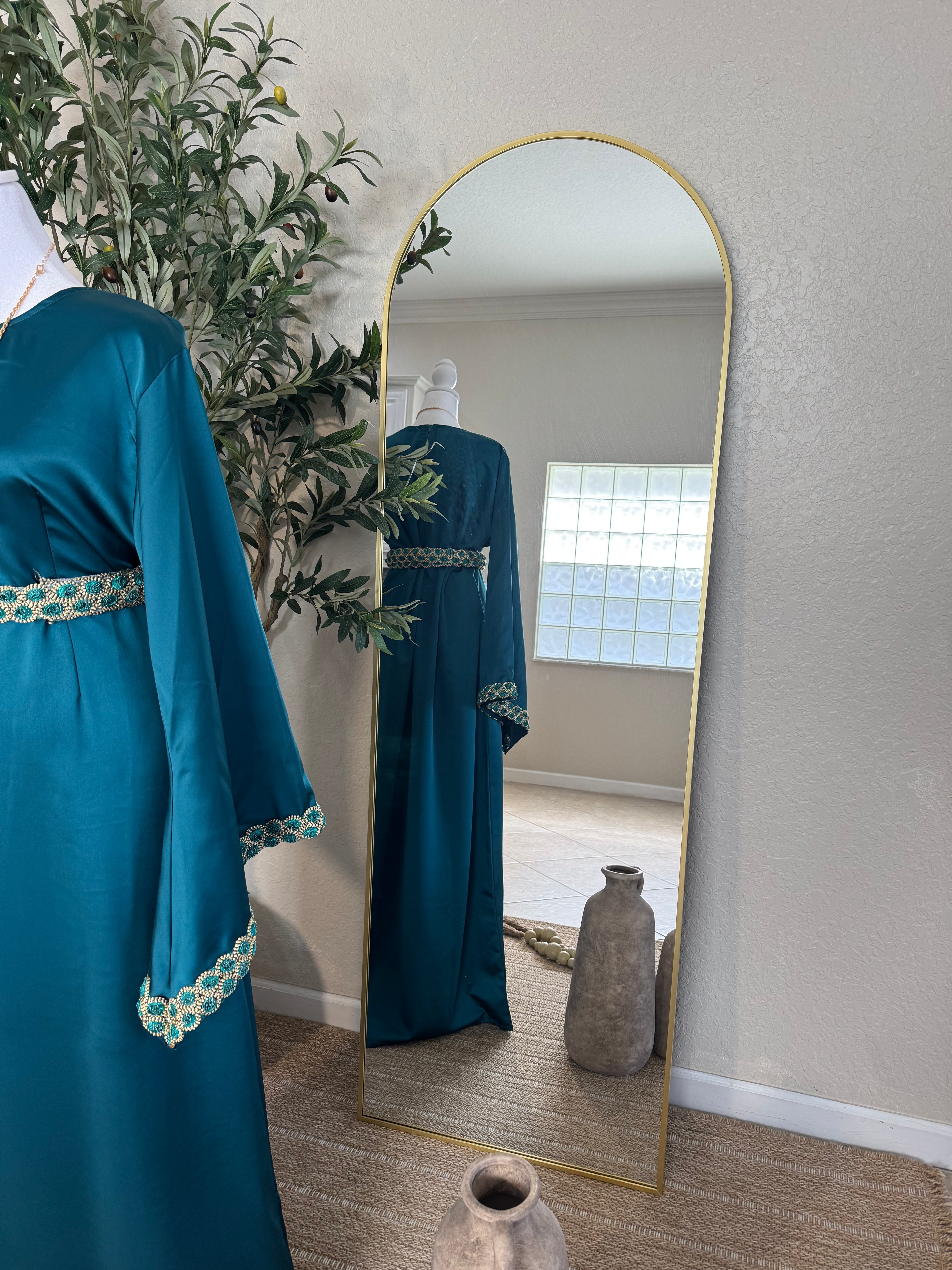 The Emerald Dress displayed on a mannequin beside a full-length mirror, featuring teal fabric with intricate gold embroidery on the sleeves and waistband, styled in a minimalist room with natural light and decorative plants.