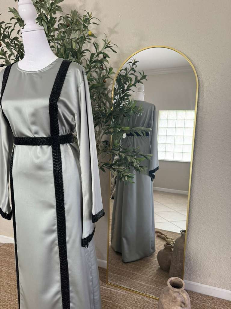 The Lunar Abaya displayed on a mannequin next to a gold-framed mirror, featuring a sleek silver fabric with black embroidered modest detailing.