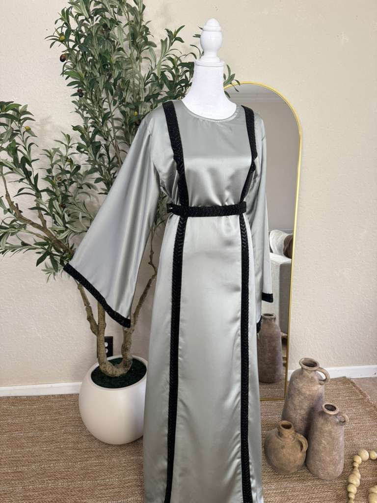 The Lunar Abaya displayed on a mannequin features a sleek silver satin fabric with elegant black embroidered trim along the sleeves, front, and belt, showcasing luxury modest fashion.