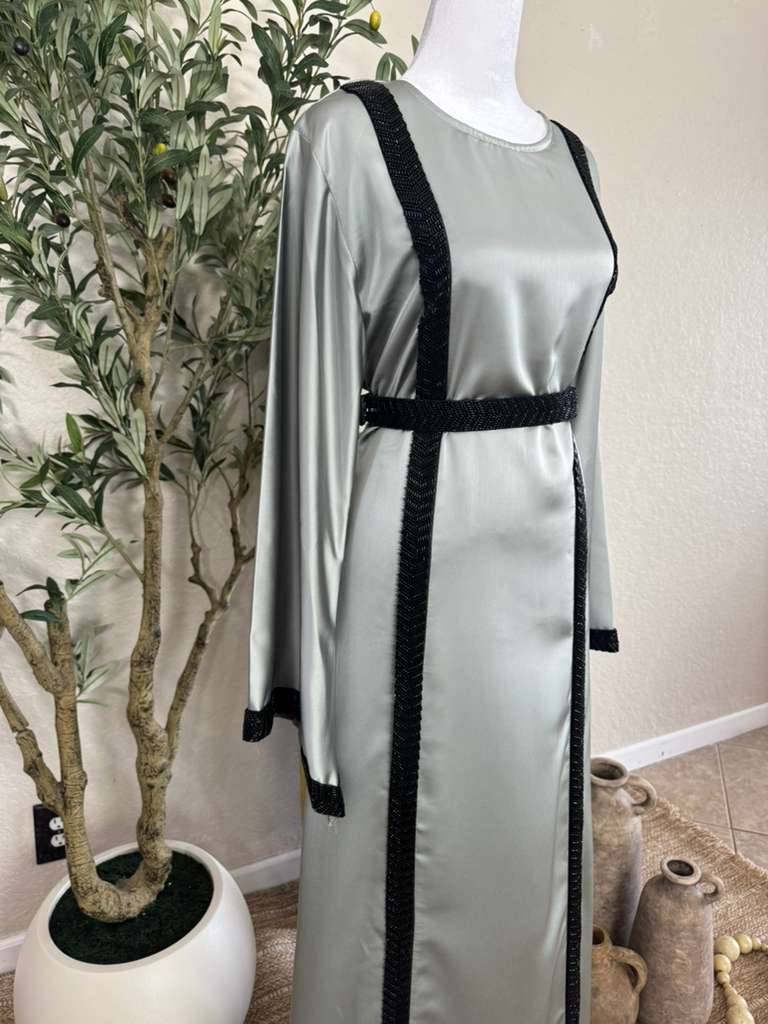 The Lunar Abaya displayed on a mannequin in a softly lit room, featuring a smooth silver fabric with bold black embroidered trim along the sleeves, front, and waist, embodying luxurious modest fashion.