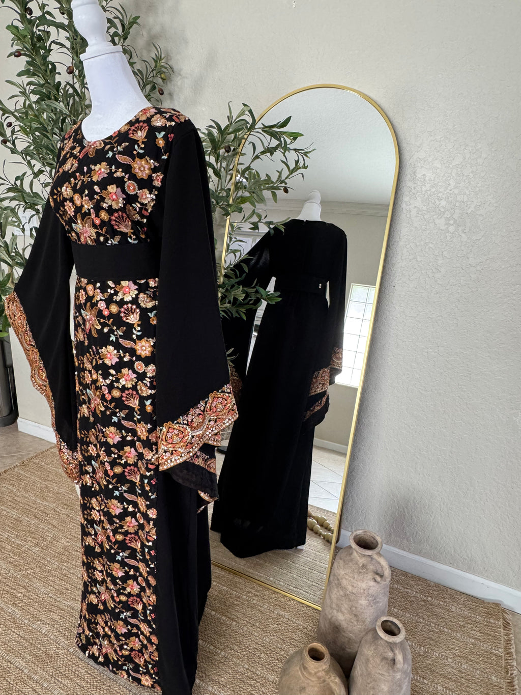 The Midnight Garden Abaya displayed on a mannequin, featuring intricate floral embroidery on black fabric with wide sleeves and detailed cuffs, styled alongside a large standing mirror and decorative vases.