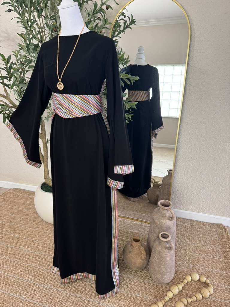 Mannequin dressed in The Olive Nights Abaya, a black modest dress featuring colorful striped detailing on the waistband, sleeves, and hem, styled with a gold pendant necklace.