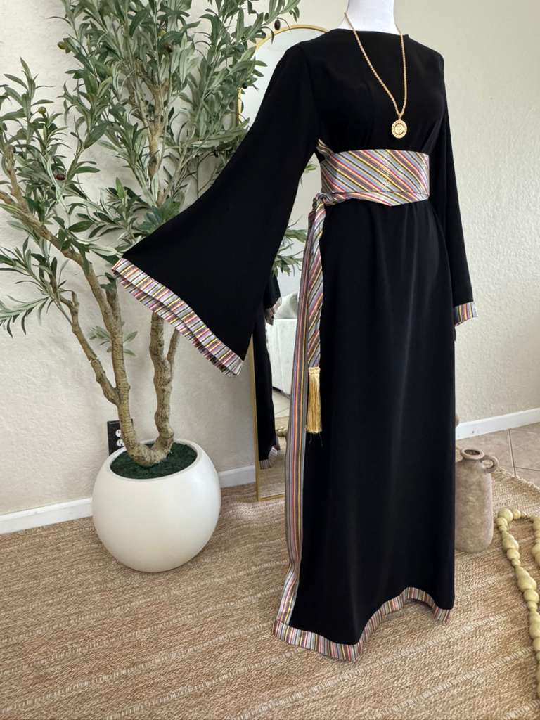 The Olive Nights Abaya displayed on a mannequin in a bright room, featuring black fabric with colorful striped accents on wide sleeves, hem, and waist belt, paired with a gold pendant necklace.