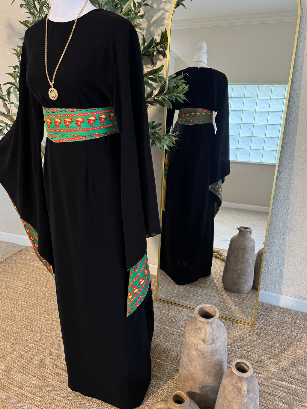 The Traditional Asba Dress displayed on a mannequin features a black modest abaya with wide sleeves and vibrant green, red, and orange embroidered accents on the waist and sleeves, styled with a gold pendant necklace.