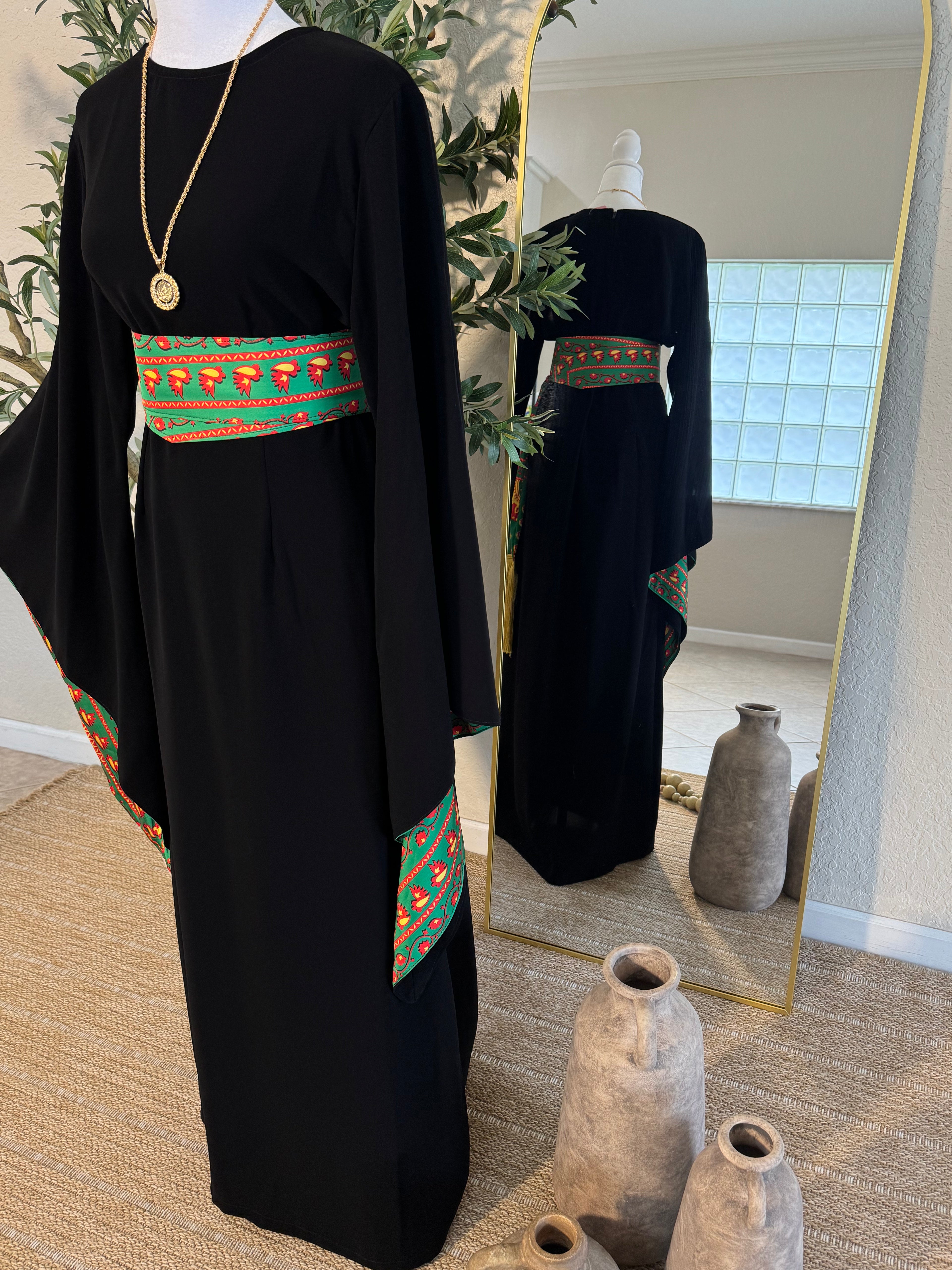 The Traditional Asba Dress displayed on a mannequin features a black modest abaya with wide sleeves and vibrant green, red, and orange embroidered accents on the waist and sleeves, styled with a gold pendant necklace.