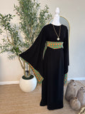The Traditional Asba Dress displayed on a mannequin features flowing black fabric with wide sleeves and colorful traditional Palestinian embroidery on the sleeves and waist, styled with a pendant necklace in a minimalist indoor setting.