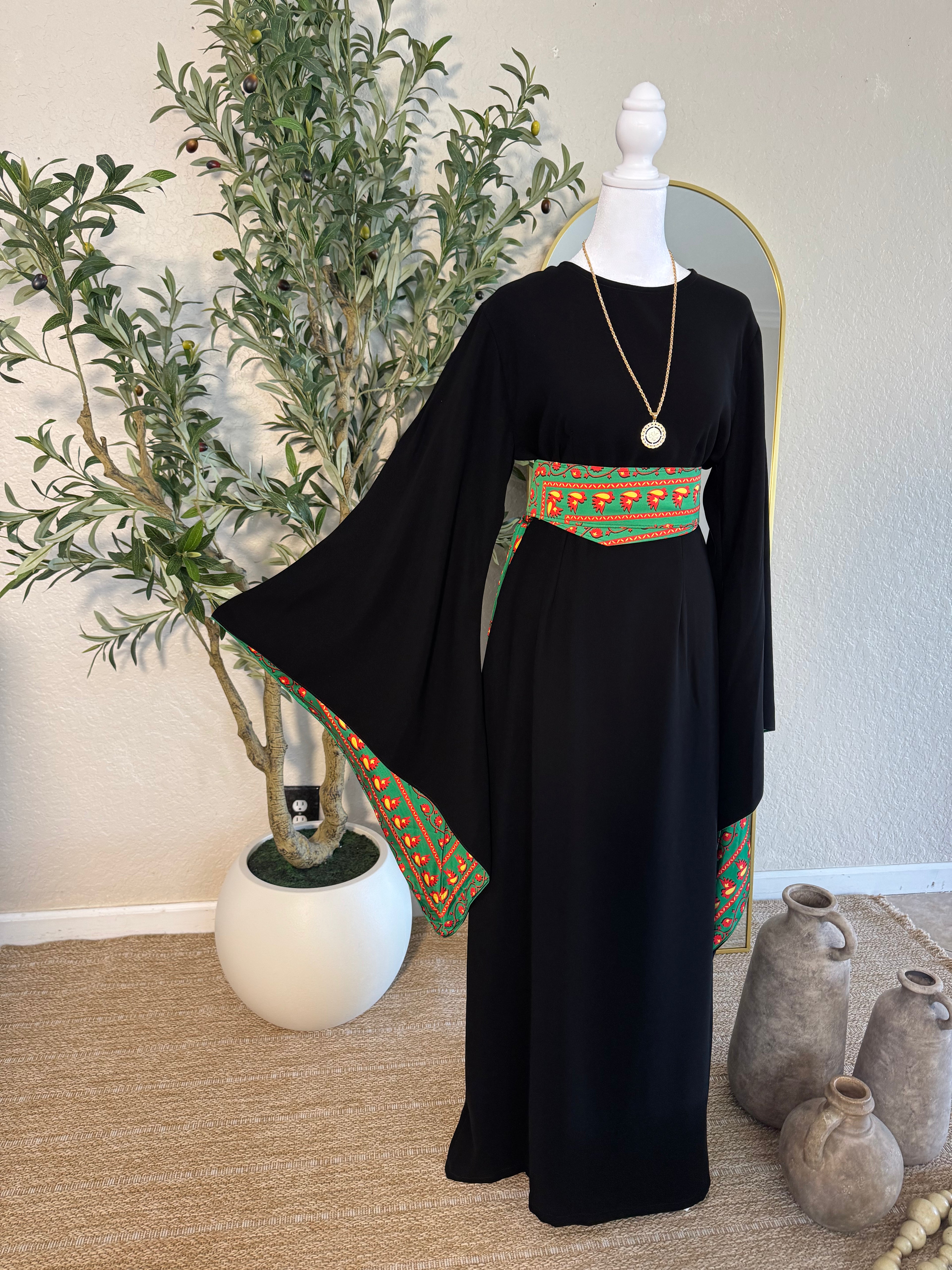 The Traditional Asba Dress displayed on a mannequin features flowing black fabric with wide sleeves and colorful traditional Palestinian embroidery on the sleeves and waist, styled with a pendant necklace in a minimalist indoor setting.