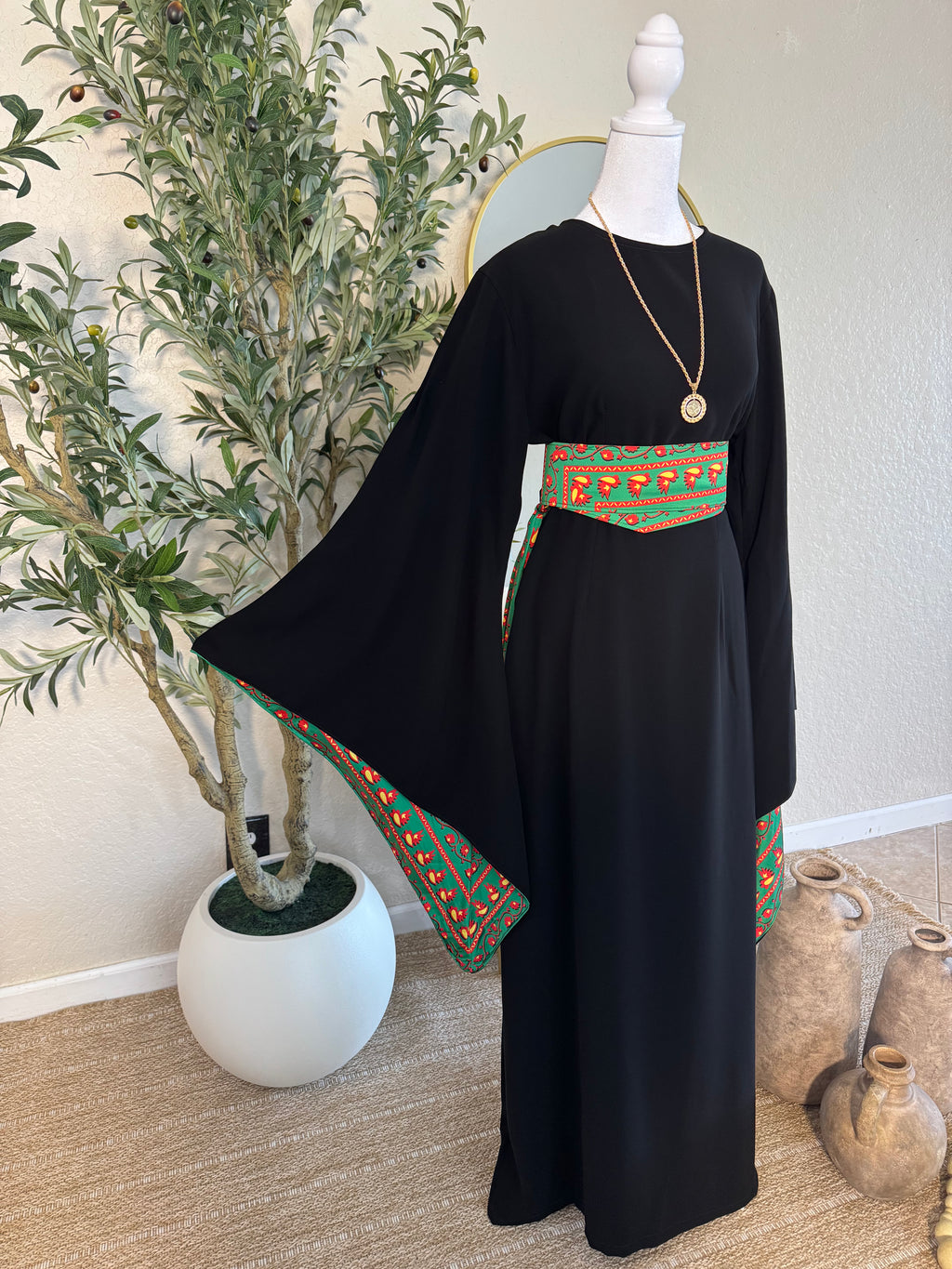 A black modest dress featuring wide sleeves and a vibrant green and red embroidered belt and sleeve trim, styled with a gold pendant necklace on a mannequin, showcasing The Traditional Asba Dress.