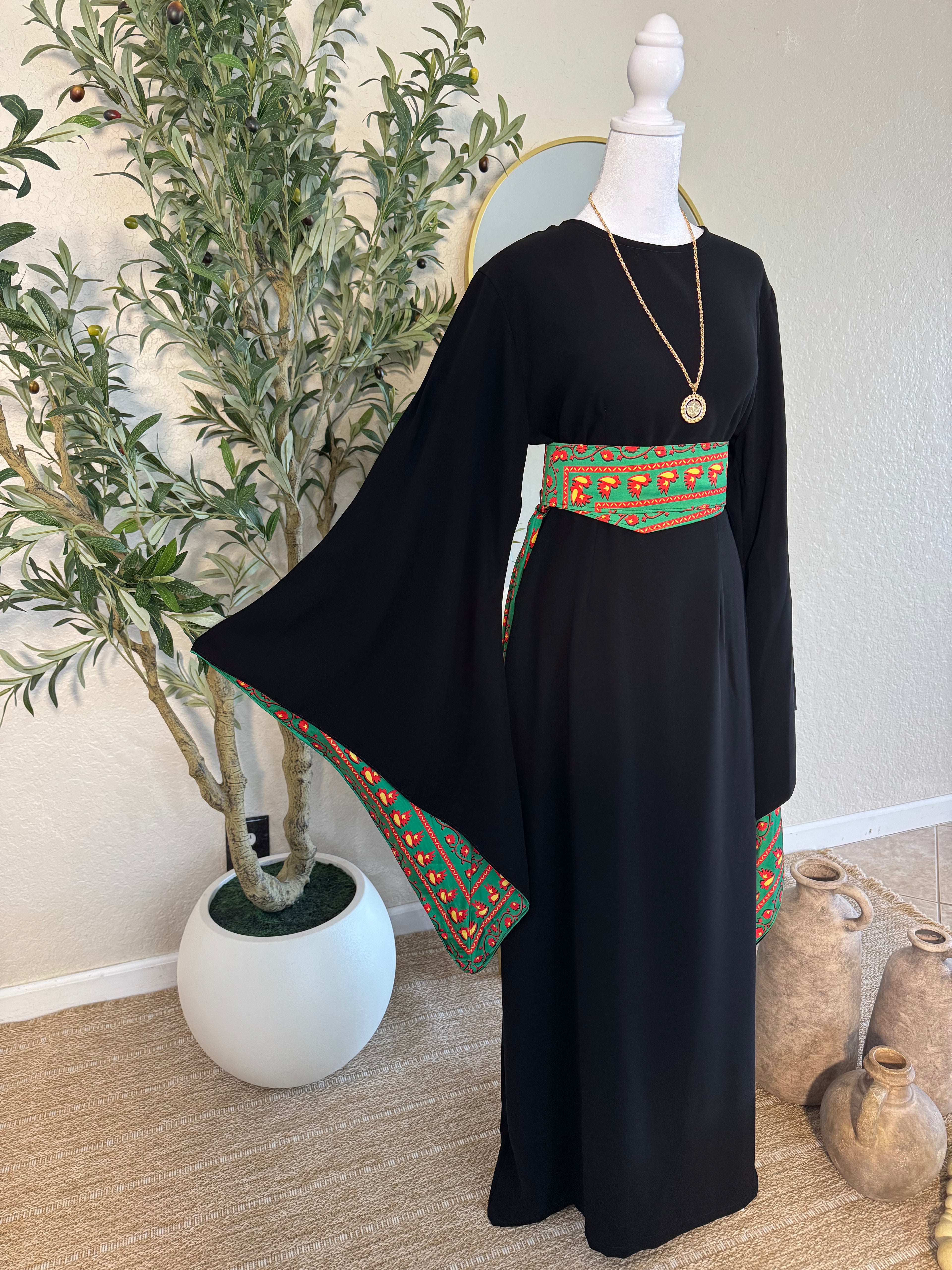 A black modest dress featuring wide sleeves and a vibrant green and red embroidered belt and sleeve trim, styled with a gold pendant necklace on a mannequin, showcasing The Traditional Asba Dress.