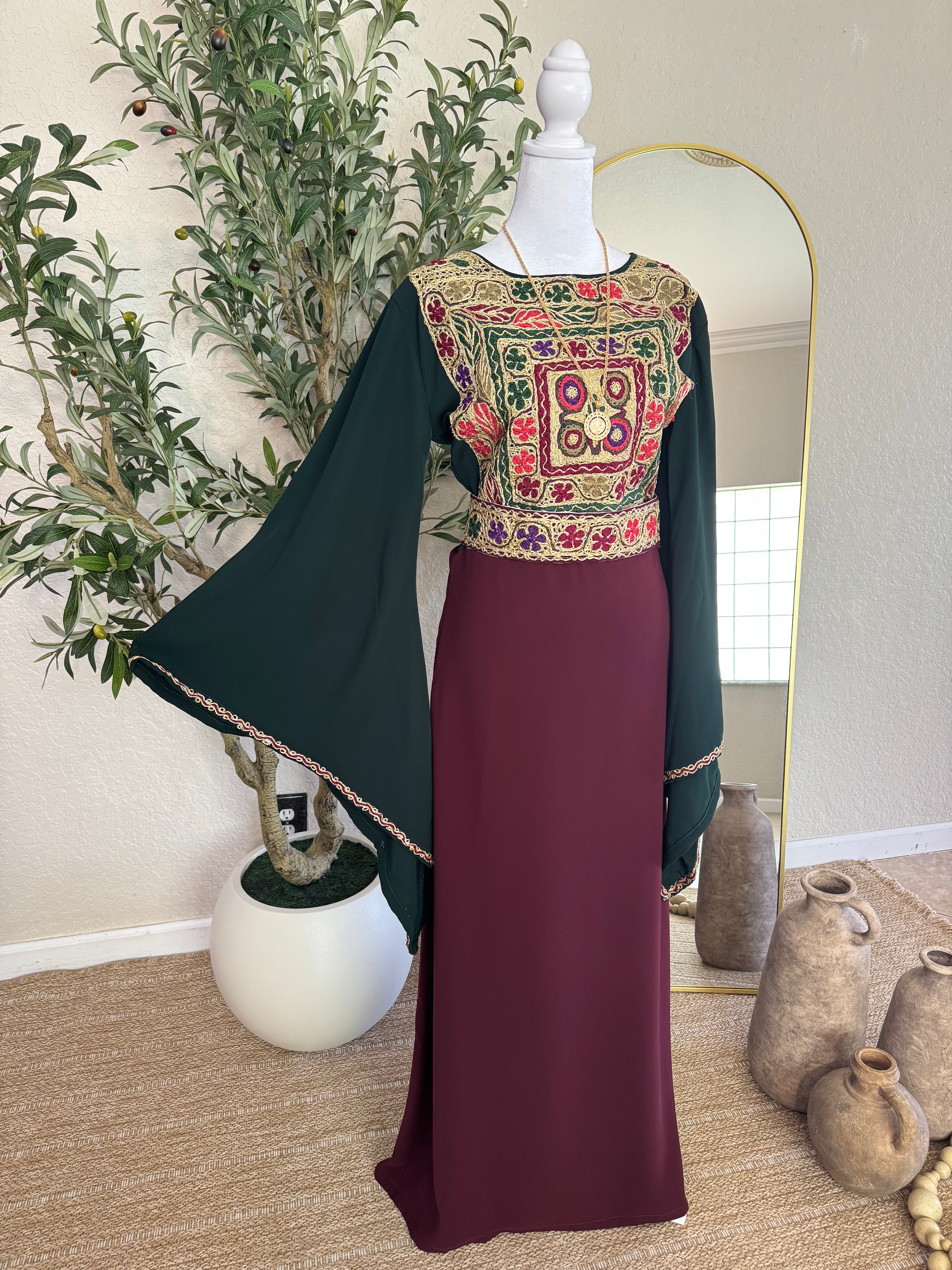 The Traditional Malaka Thoub displayed on a mannequin featuring intricate multicolor floral embroidery on the bodice, black flowing sleeves with detailed trim, and a deep burgundy floor-length skirt, set against a neutral background with decorative vases and greenery.