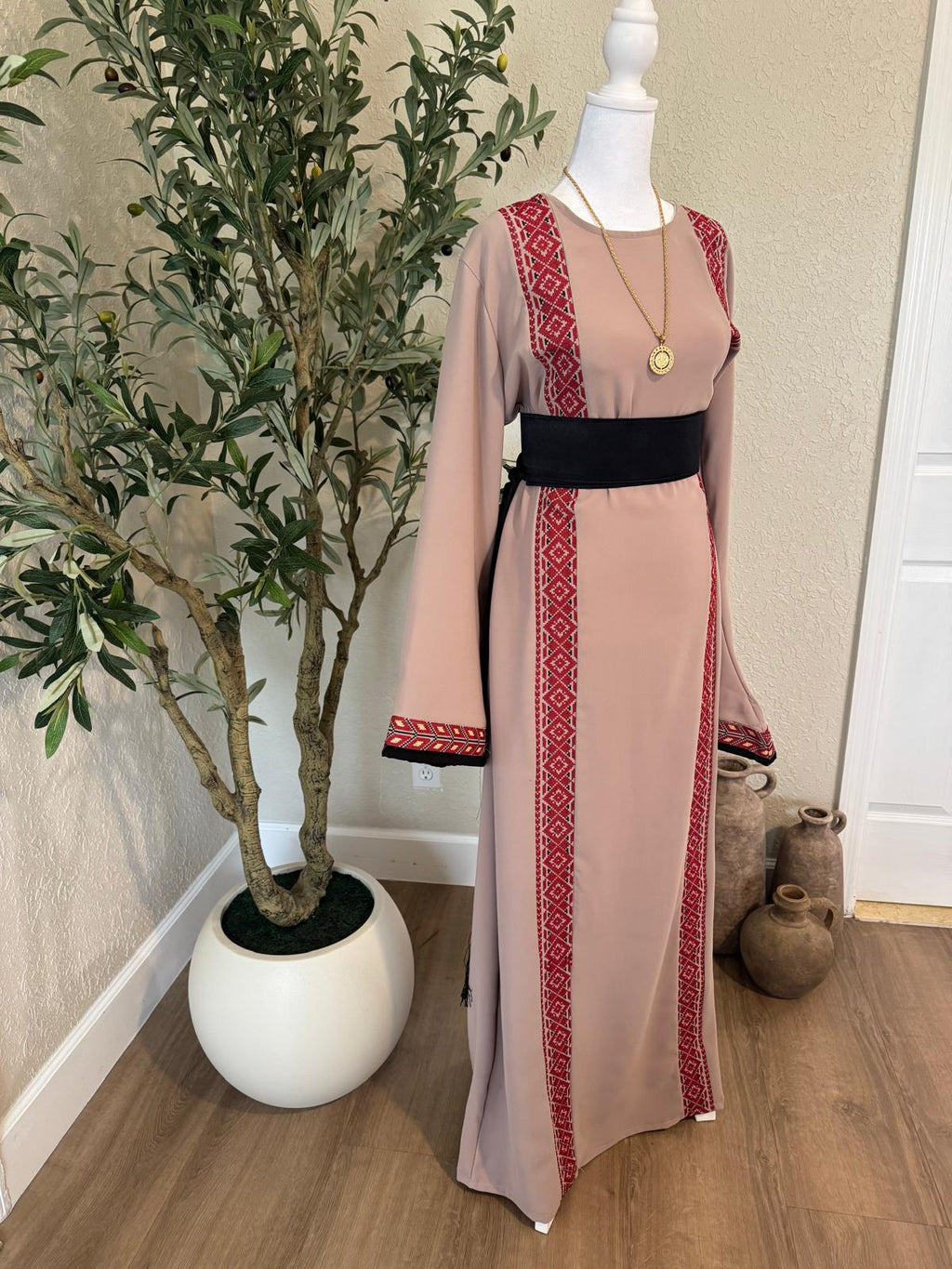 Front view of The Ramallah Line Palestinian Abaya in dusty taupe with red geometric vertical detailing and black waist belt