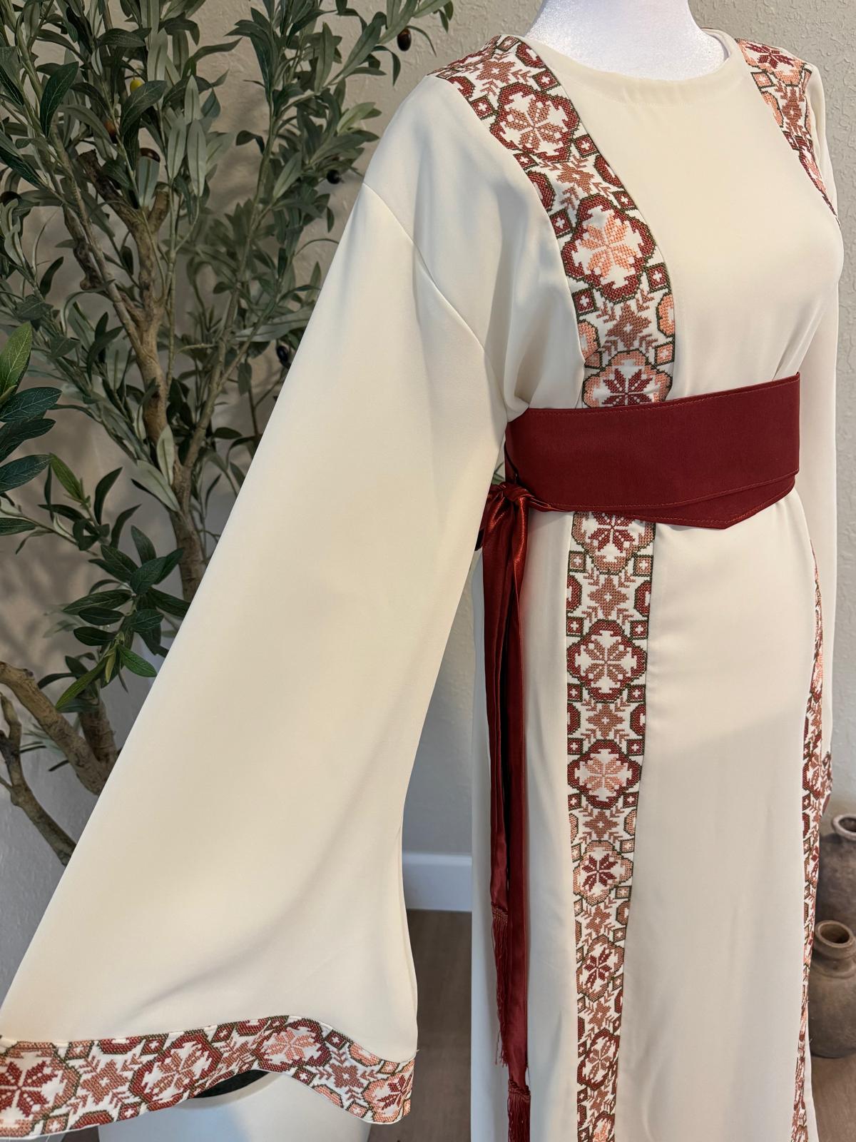 Full-length view of The: Bethlehem Sand Thobe with ivory fabric, vertical terracotta patterned panels, flowing sleeves and rust-red belt